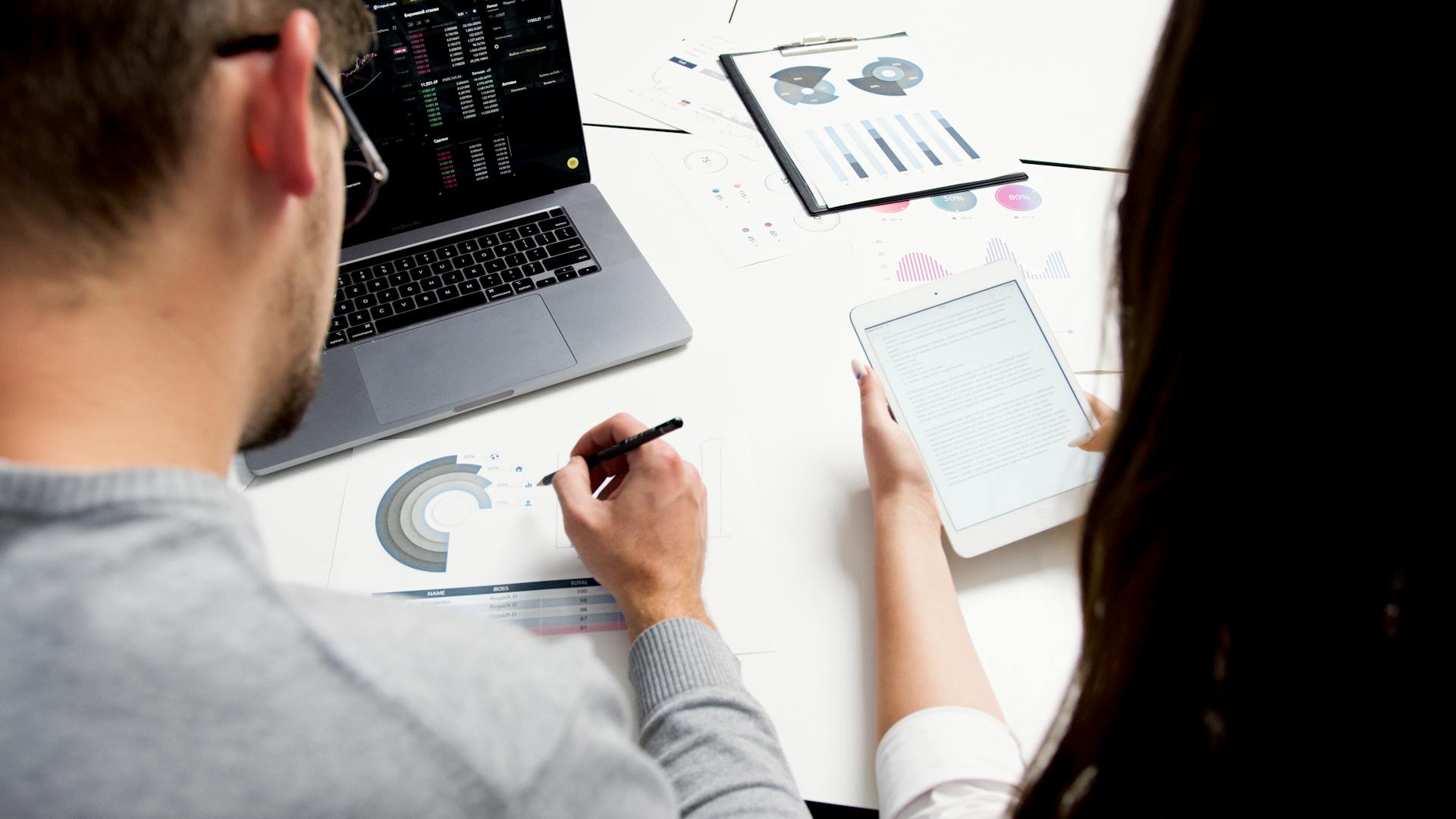 Two colleagues reviewing charts on a laptop, a tablet and printed reports at a desk.