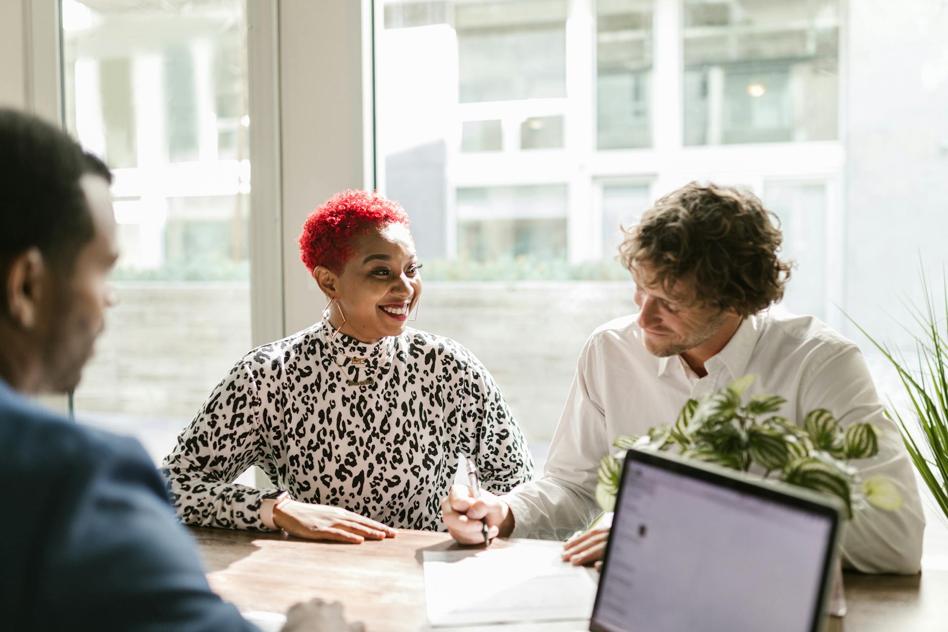 Three colleagues talking at a table with a laptop by a bright window.